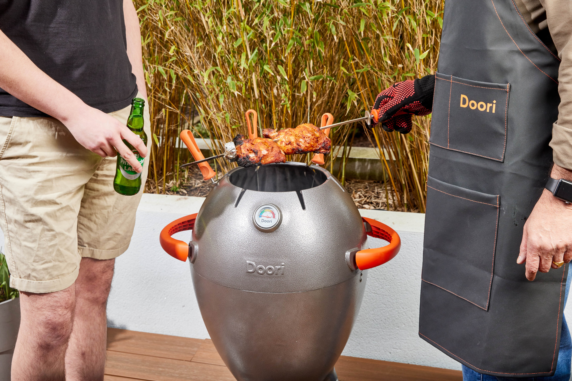 Two men stood over the Doori Outdoor Cooker, one holding a beer and one wearing the Doori apron and gloves and holding a skewer of chicken.