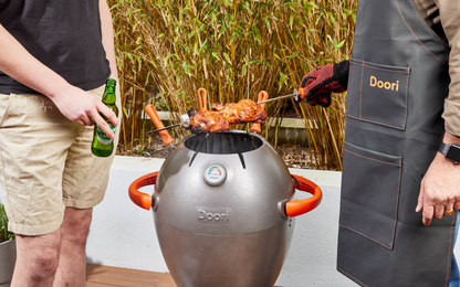 Two men stood over the Doori Outdoor Cooker, one holding a beer and one wearing the Doori apron and gloves and holding a skewer of chicken.