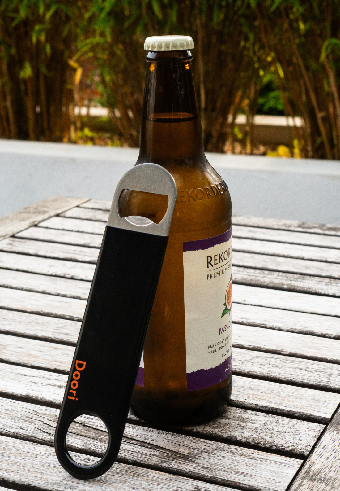 The Doori Bottle Opener leaning against a bottle of cider on an outdoor table.